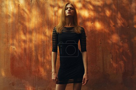 Little black dress concept. Emotive portrait of a proud fashionable model posing over rusty stone wall with plashes of sunlight on it. Ultra fashion style. Copy-space. Outdoor shot