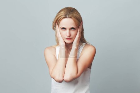 Close-up portrait of a young woman in white sleeveless shirt imitating hear no evil concept on gray background. Human emotions, expressions, communication. Text space. Studio shot