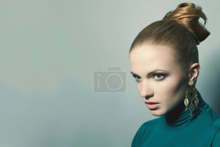Photo for Female fashion concept. Portrait of beautiful young blonde wearing emerald green dress, ethnic earrings. Model posing over light-blue background. Perfect skin, stylish hairdo. Close up. Copy-space. Studio shot - Royalty Free Image