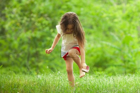 Photo for After rain concept. Stylish baby girl with long brown hair in vintage red and white dress standing in the park and trying to put on a shoe. Sunny weather. Text space. Outdoor shot - Royalty Free Image