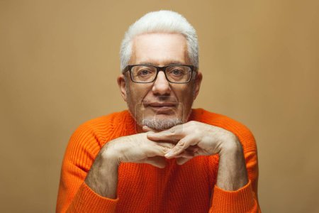 Téléchargez les photos : Fabuleux à tout âge, concept de lunettes. Portrait d'homme à la mode âgé de 60 ans en pull orange sur fond beige pastel. Superbe coupe de cheveux, cheveux gris brillant. Poignée poignet-serrure des mains. Espace de copie - en image libre de droit