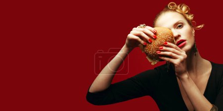Photo for Fahion Fast Food Concept. Guilty pleasure. Fashionable model holding burger near face over red background. Perfect hair, skin, make-up, manicure. Golden accessories. Close up. Copy-space. Studio shot - Royalty Free Image