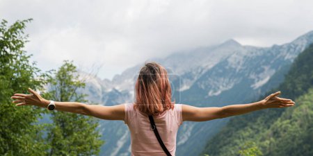 Vista desde atrás de una joven con los brazos extendidos afuera rodeada de montañas y naturaleza.