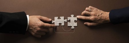Wide view image of businessman and businesswoman hands joining two blank matching puzzle pieces. Over brown background.