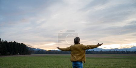 Foto de Hombre joven en chaqueta de color ocre mindfully de pie en la hermosa naturaleza verde bajo el cielo nublado de la noche con sus brazos abiertos. - Imagen libre de derechos