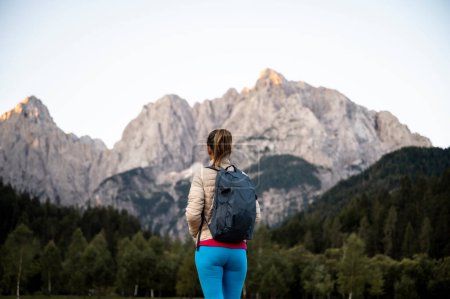 Foto de Vista desde atrás de una excursionista femenina con mochila de pie y mirando una hermosa vista de las altas montañas. - Imagen libre de derechos