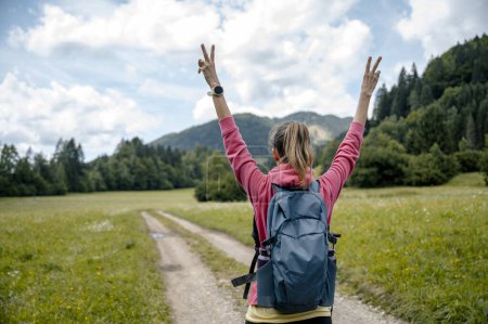 Foto de Vista desde atrás de una excursionista femenina con mochila en la espalda de pie en un camino de hermosa naturaleza verde con los brazos levantados en alto en gesto victorioso. - Imagen libre de derechos