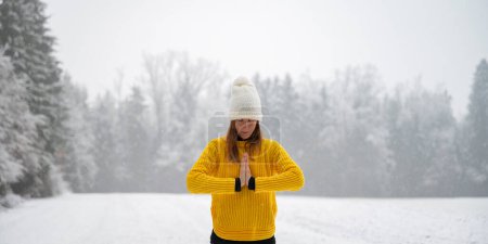 Foto de Mujer joven pacífica en suéter amarillo y sombrero blanco meditando o rezando con sus palmas unidas frente a su pecho mientras está de pie afuera en la naturaleza nevada del invierno. - Imagen libre de derechos