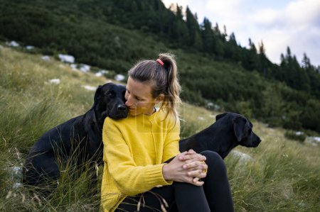 Foto de Joven propietaria de dos perras labradoras negras de raza pura beautfiul sentadas en hierba verde en la colina con uno de sus perros apoyando la cabeza sobre el hombro de las mujeres en un gesto amoroso. - Imagen libre de derechos