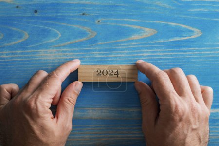 Foto de Vista aérea de manos masculinas colocando etiqueta de madera con signo 2024 sobre fondo de madera azul texturizado. - Imagen libre de derechos