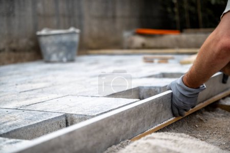 Foto de Vista de ángulo bajo de manos masculinas midiendo y nivelando baldosas de arena y cemento para un pavimento preciso del patio exterior o acera. Industria de la construcción y renovación. - Imagen libre de derechos