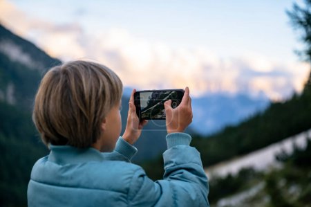 Foto de Por encima de la vista del hombro de un niño pequeño tomando una foto con su teléfono inteligente de hermosas montañas mientras toma un descanso durante el senderismo. - Imagen libre de derechos