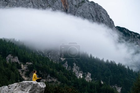 Foto de Joven mujer serena sentada en posición de loto sobre una roca en las altas montañas y meditando. Con suave niebla de niebla o nubes pasando por la hermosa naturaleza. - Imagen libre de derechos