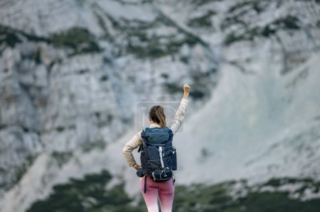 Foto de Vista desde atrás de una joven excursionista victoriosa con una mochila de pie en las altas montañas con el brazo en alto. - Imagen libre de derechos