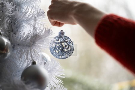Foto de Vista de cerca de la mano femenina en suéter rojo colgando adorno de vacaciones azul brillante en un árbol de Navidad blanco. - Imagen libre de derechos