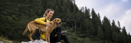 Foto de Amplia imagen de una joven mujer descansando sobre una roca durante una caminata con su hermoso perro perro perro perro perdiguero dorado junto a ella. - Imagen libre de derechos