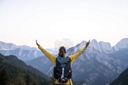 Foto de Vista desde atrás de una excursionista femenina con una mochila de pie con los brazos extendidos mirando una hermosa vista de las altas montañas bajo el cielo despejado. - Imagen libre de derechos