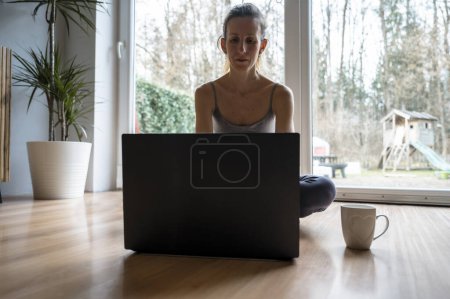 Foto de Mujer joven, freelancer, sentada en el suelo de su sala de estar trabajando en un ordenador portátil con una taza de café a su lado. - Imagen libre de derechos
