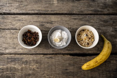 Foto de Vista superior de tres tazones con ingredientes saludables para el desayuno: pasas de uva, yogur y granola con un plátano maduro a su lado. Sobre tableros de madera rústicos texturizados. - Imagen libre de derechos