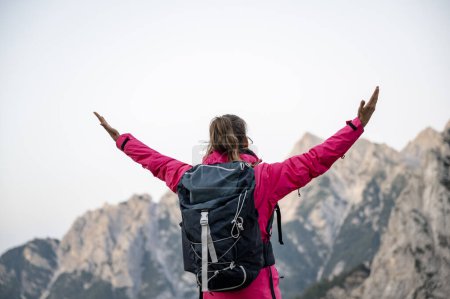 Foto de Joven excursionista en chaqueta rosa y mochila en sus hombros atnding con sus brazos elevados en celebración como ella mira hermosas montañas altas. - Imagen libre de derechos