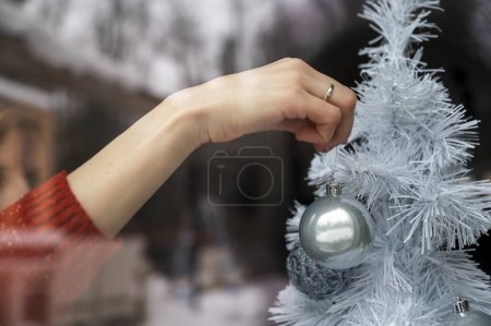 Foto de Vista a través de la ventana de una mujer que cuelga adorno azul brillante del día de fiesta en un árbol de Navidad blanco. Con la naturaleza nevada de invierno reflejándose en el vaso. - Imagen libre de derechos