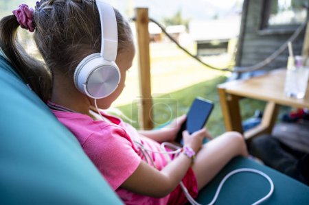 Foto de Vista lateral sobre el hombro de una niña pequeña sentada en un café con auriculares en los oídos jugando juegos o escuchando música en el teléfono móvil. - Imagen libre de derechos