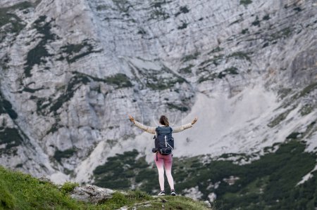 Foto de Vista desde atrás de una joven excursionista con una mochila de pie en la cima de una montaña con los brazos extendidos en celebración mientras mira una vista de otra alta montaña rocosa. - Imagen libre de derechos