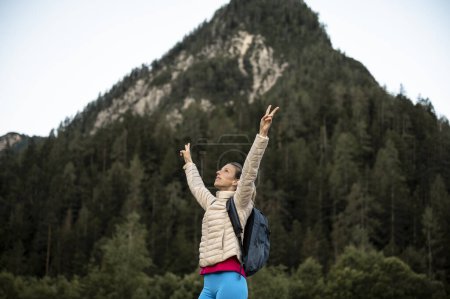 Foto de Mujer joven, una excursionista con mochila, de pie con los brazos en alto con gesto victorioso fuera en la naturaleza montañosa. - Imagen libre de derechos
