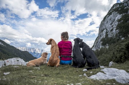 Foto de Vista desde atrás de una mujer joven y sus cuatro perros sentados en un pico de montaña mirando a la hermosa vista de la naturaleza con el cielo nublado blanco. - Imagen libre de derechos