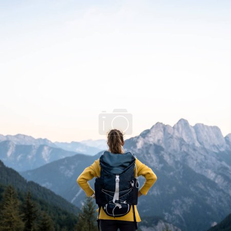 Foto de Vista desde atrás de una excursionista femenina con mochila de pie en la cima de una montaña mirando una hermosa vista de otros picos altos. Bajo cielo despejado. - Imagen libre de derechos