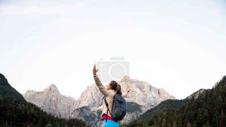 Foto de Feliz joven excursionista de pie rodeada de hermosas montañas altas con los brazos extendidos mientras disfruta de su victoria en el senderismo. - Imagen libre de derechos