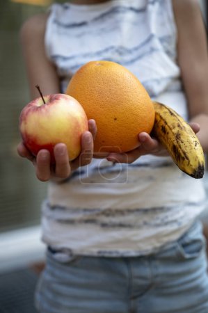 Foto de Vista de cerca de un niño sosteniendo tres pedazos de fruta - manzana, naranja y plátano - en sus manos. - Imagen libre de derechos
