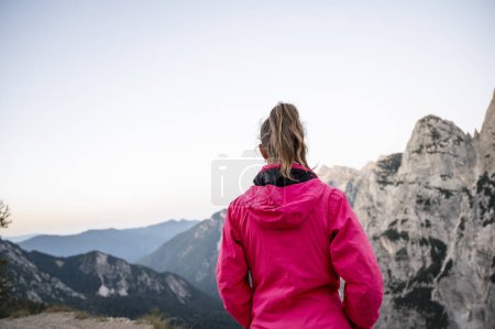 Foto de Vista desde atrás de una excursionista femenina con chaqueta rosa de pie mirando a la hermosa vista de la montaña. Paz y soledad imagen conceptual. - Imagen libre de derechos