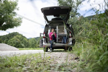 Foto de Mujer joven en un viaje en carretera con sus perros descansando mientras se sienta en un baúl abierto de su coche lleno de cajas de perro y navega por el teléfono móvil para obtener direcciones e información sobre el destino. - Imagen libre de derechos