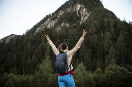 Foto de Joven excursionista con una mochila de pie bajo una montaña con los brazos levantados en alto en gesto victorioso. Imagen conceptual de la esperanza, el futuro y la creencia. - Imagen libre de derechos