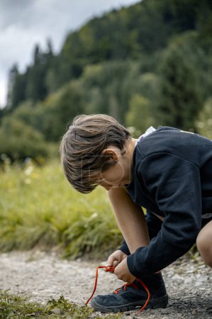Foto de Niño arrodillado en un sendero en la naturaleza verde para atar sus zapatos de senderismo en un lazo. Tiempo de vacaciones y aventura al aire libre. - Imagen libre de derechos