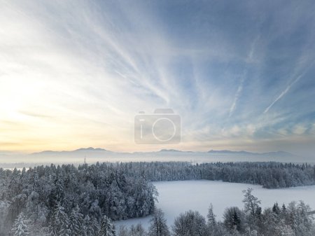 Foto de Foto con un dron de un hermoso paisaje cubierto de nieve de invierno - bosque y un claro con montañas en la distancia bajo el hermoso cielo azul con suaves couds de niebla. - Imagen libre de derechos
