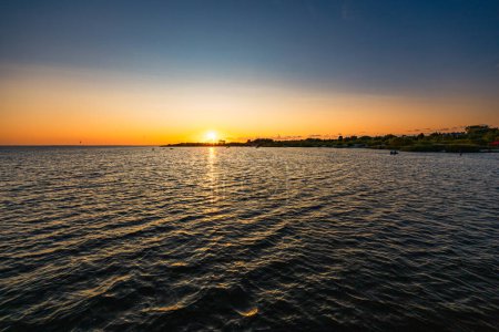 Téléchargez les photos : Beau coucher de soleil nuageux sur la mer avec quelques bateaux et kitesurfers et quelques arbres et buissons à côté de la petite plage - en image libre de droit