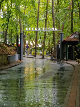 Photo for Sopot, Poland - July 23 2023: Gate on the road as entrance to Opera Lesna in Sopot city in forest with glowing lamp with name reflecting in road wet from rain - Royalty Free Image