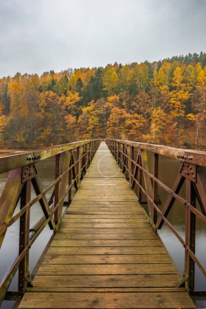 Beautiful view to long steel and wood bridge over big and silent lake with autumn golden trees and bushes around at cloudy and rainy morning