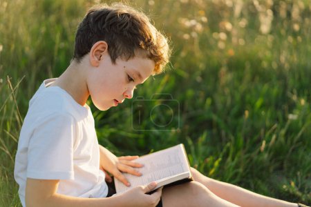 Christlicher Junge hält Bibel in ihren Händen. Lesen der Heiligen Bibel auf einem Feld bei schönem Sonnenuntergang. Konzept für Glauben, Spiritualität und Religion. Frieden, Hoffnung