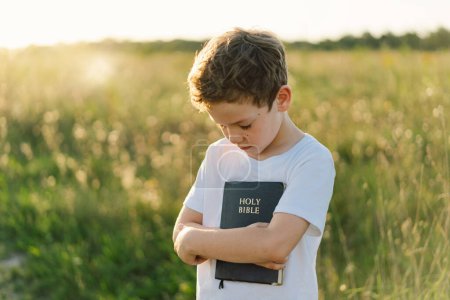 Christian Boy tient la bible dans ses mains. Lire la Sainte Bible dans un champ pendant un beau coucher de soleil. Concept de foi, de spiritualité et de religion. Paix, espérance