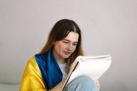 A Ukrainian teenage girl is wrapped in the Ukrainian flag and is drawing or doing homework in the bedroom. A teenager studies at home. The concept of studies during the war in Ukraine.