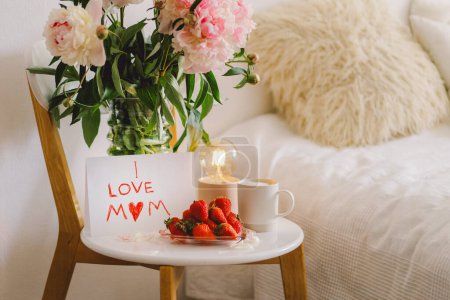 Breakfast for Mothers Day. Heart shaped white plate with fresh strawberries, cup of coffee, gift and Peonys bouquet with gift in bed. Still life composition. Happy Mothers Day.