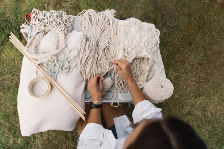 Woman knits bag using macrame technique outdoors near tents. Outdoor hobbies. Macrame weaving.