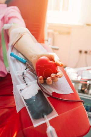 Téléchargez les photos : Une jeune femme qui fait un don de sang à l'hôpital. Une femme fait un don de sang tout en tenant un cœur rouge. Don de sang. - en image libre de droit