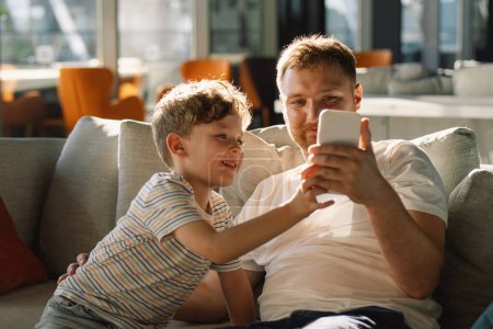 Father and son sit relax on couch in hotel hall have fun using cellphone together. Boy and dad makes selfie phone. Father and little boy child rest on sofa and watching funny video on smartphone.
