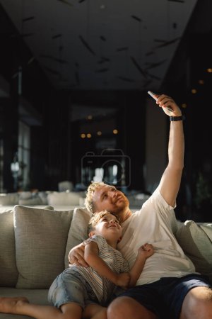 Vater und Sohn sitzen entspannt auf der Couch im Hotelflur und haben Spaß beim gemeinsamen Telefonieren. Junge und Papa machen Selfie-Telefone. Vater und kleines Kind ruhen sich auf Sofa aus und schauen lustiges Video auf Smartphone.