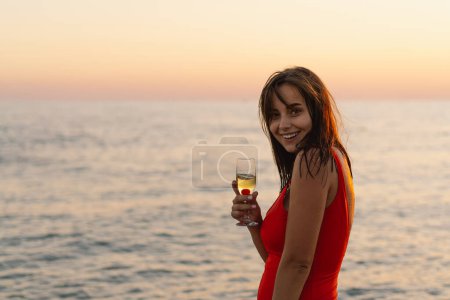 A cheerful woman in a vibrant red swimsuit stands by the seaside, holding a glass of white wine. The setting sun casts a warm golden hue over the scene, highlighting her joyous expression.