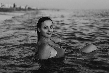 A young woman with a smile on her face is swimming in the calm ocean waters during the early evening. Her head and shoulders are visible above the waterline